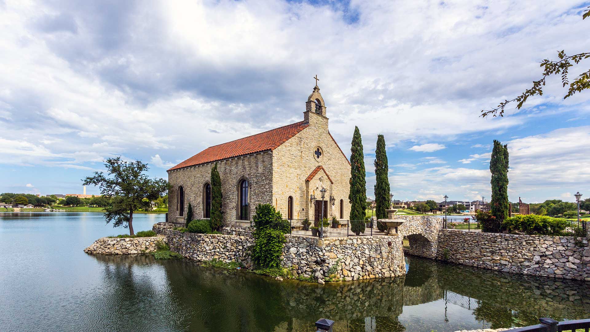 The Chapel at Adriatica sits on a stone foundation on the lake. It is a small stone structure with various plants around it.
