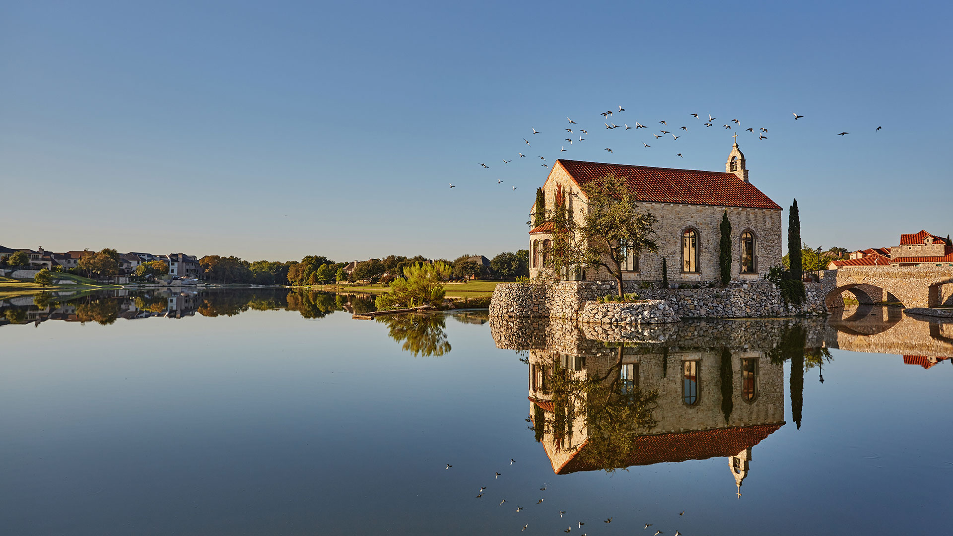 Looking across the lake at the Bella Donna Chapel at Adritica. The water is smooth, like glass and the reflection of the chapel is seen clearly. The foundation is stone as is the chapel. Trees are seen far off on the other side of the lake.