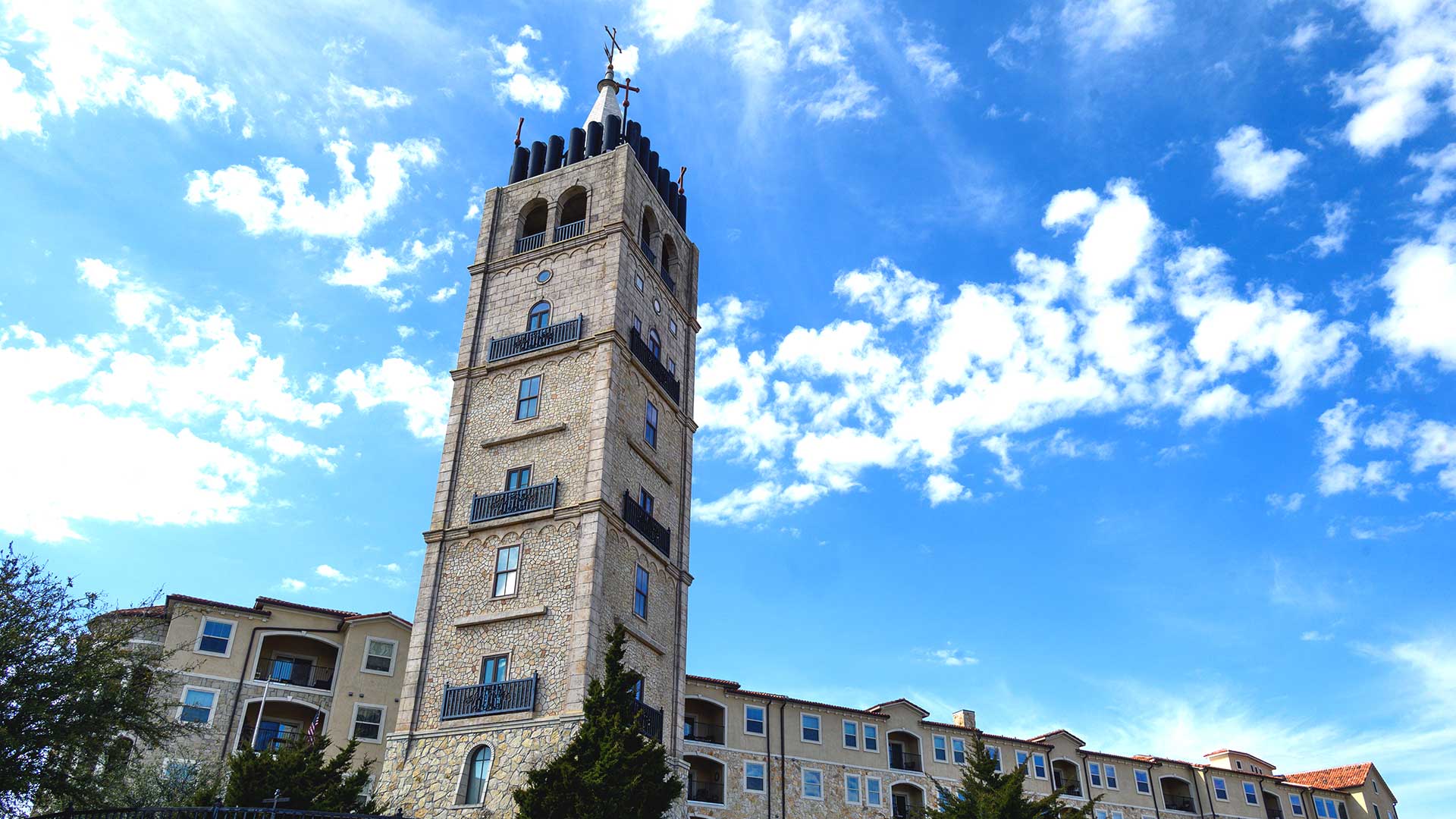 Looking up the stone Bell Tower at Adriatica. A matching apartment building is in the background on a partially cloudy day.