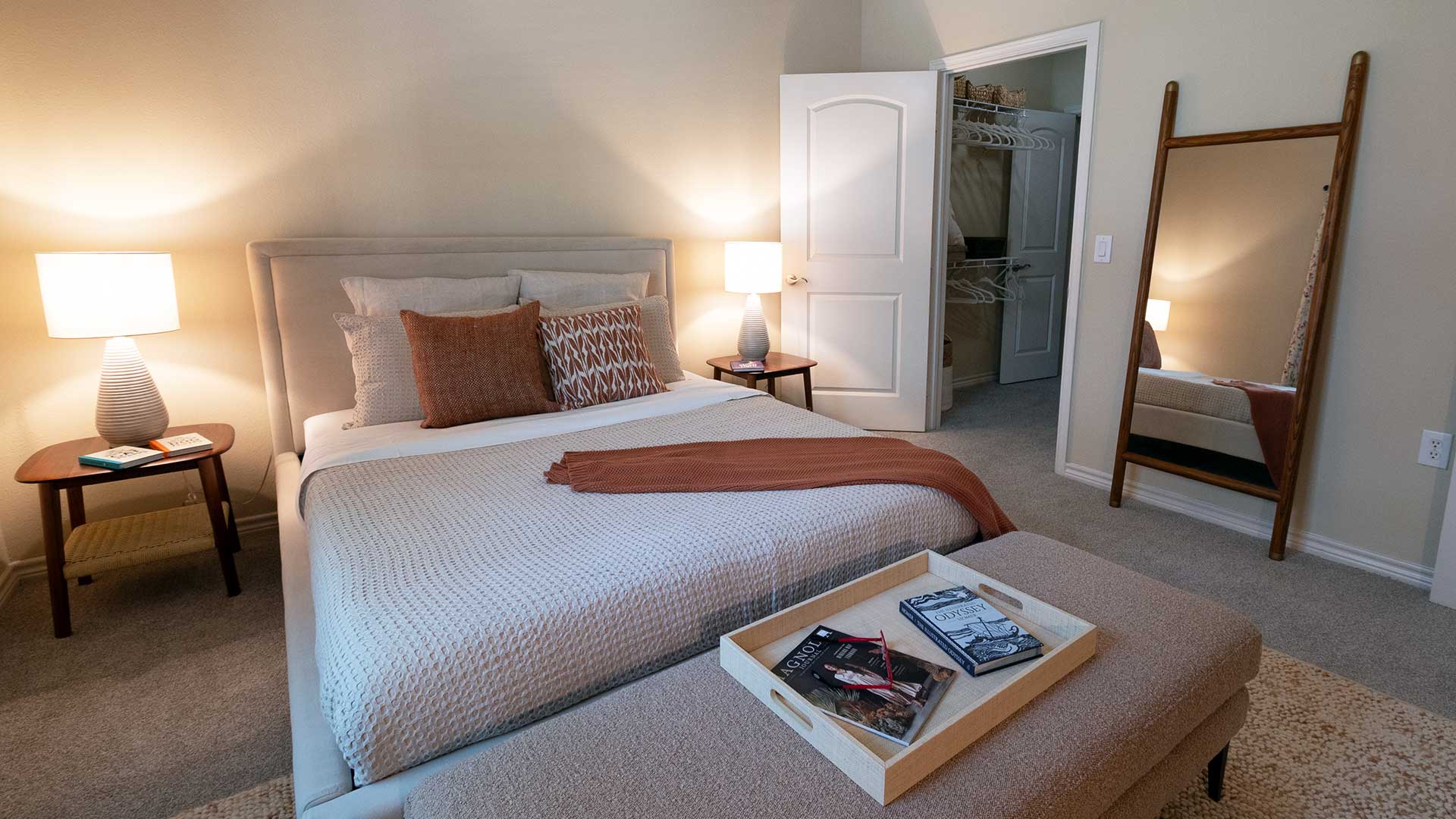 Looking across a large bed with an end table and lit lamp on either side. Behind is a doorway into a closet with a full-body mirror next to it.