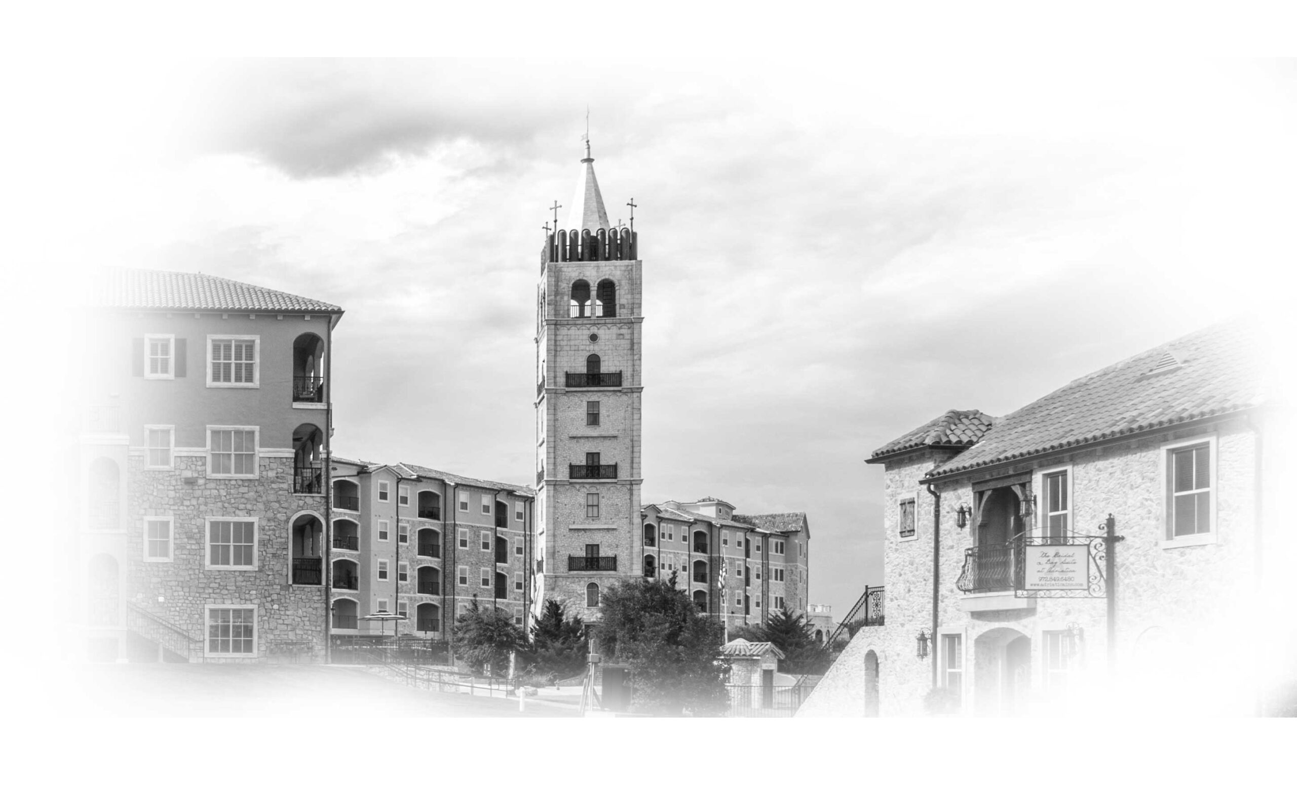 Black and white image of a Mediterranean-style residential building with stone accents and a prominent bell tower in the background, framed by a cloudy sky and additional architecture in the foreground.